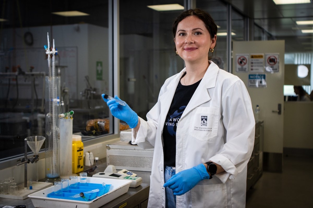 A female scientist in a white lab coat and blue gloves standing in a laboratory, holding a small vial. She is smiling and looking at the camera, with laboratory equipment and glassware on the bench beside her.