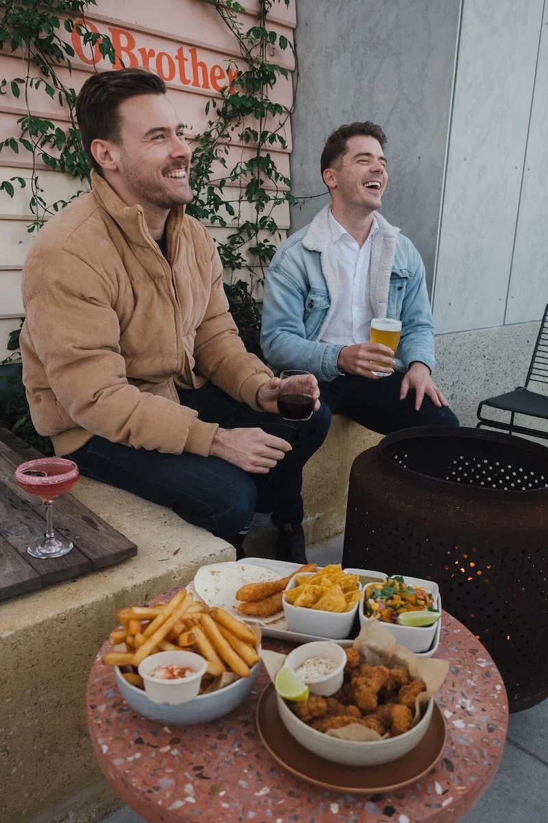 Two men sitting outdoors, smiling and enjoying drinks, with a table of various foods including french fries, chicken wings, guacamole, and drinks.