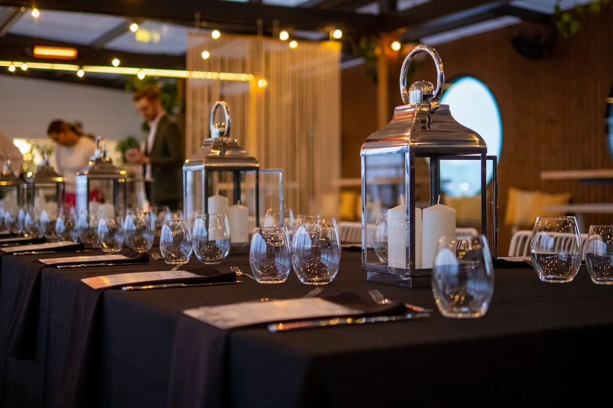 A decorated table set for a formal event with glass lanterns, candles, wine glasses, and cutlery, in a warmly lit indoor or outdoor space.