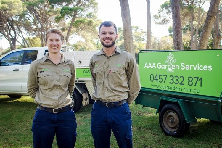 Two AAA Garden Services employees in uniform standing outdoors near a green company sign and vehicle, with trees in the background.
