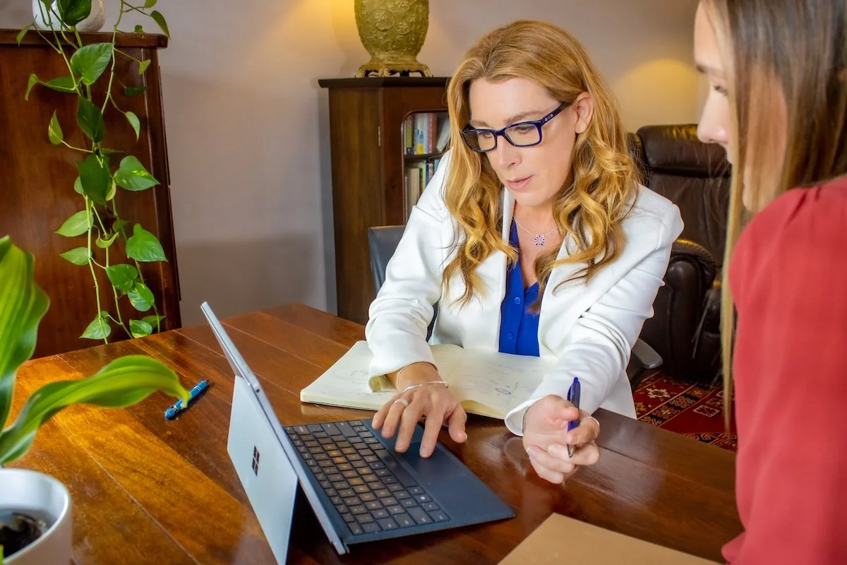 Two women sitting at a wooden table, engaged in a discussion. One woman has red hair, glasses, and is taking notes, while the other woman has brown hair and is listening. A laptop, a notebook, and a pen are on the table. There are bookshelves, plants