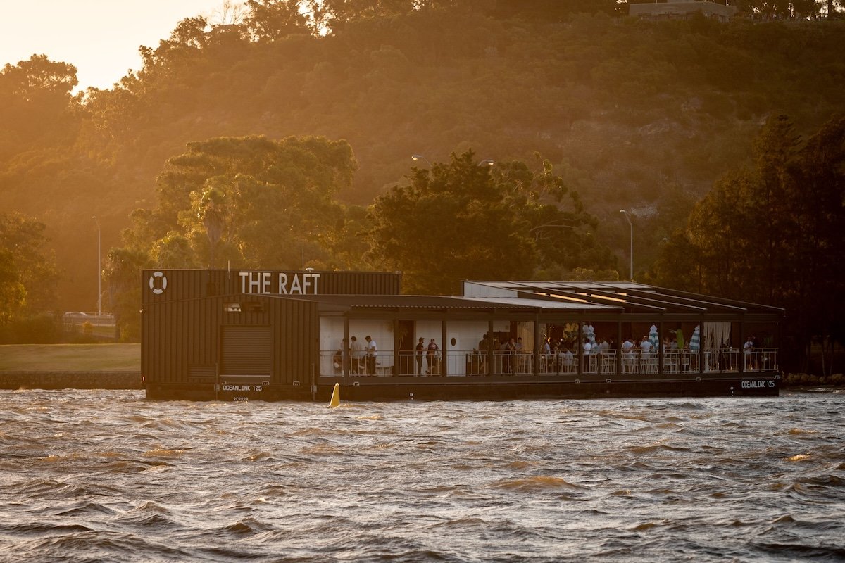A floating restaurant named 'The Raft' on a river during sunset with people dining on the deck, with trees and hills in the background.