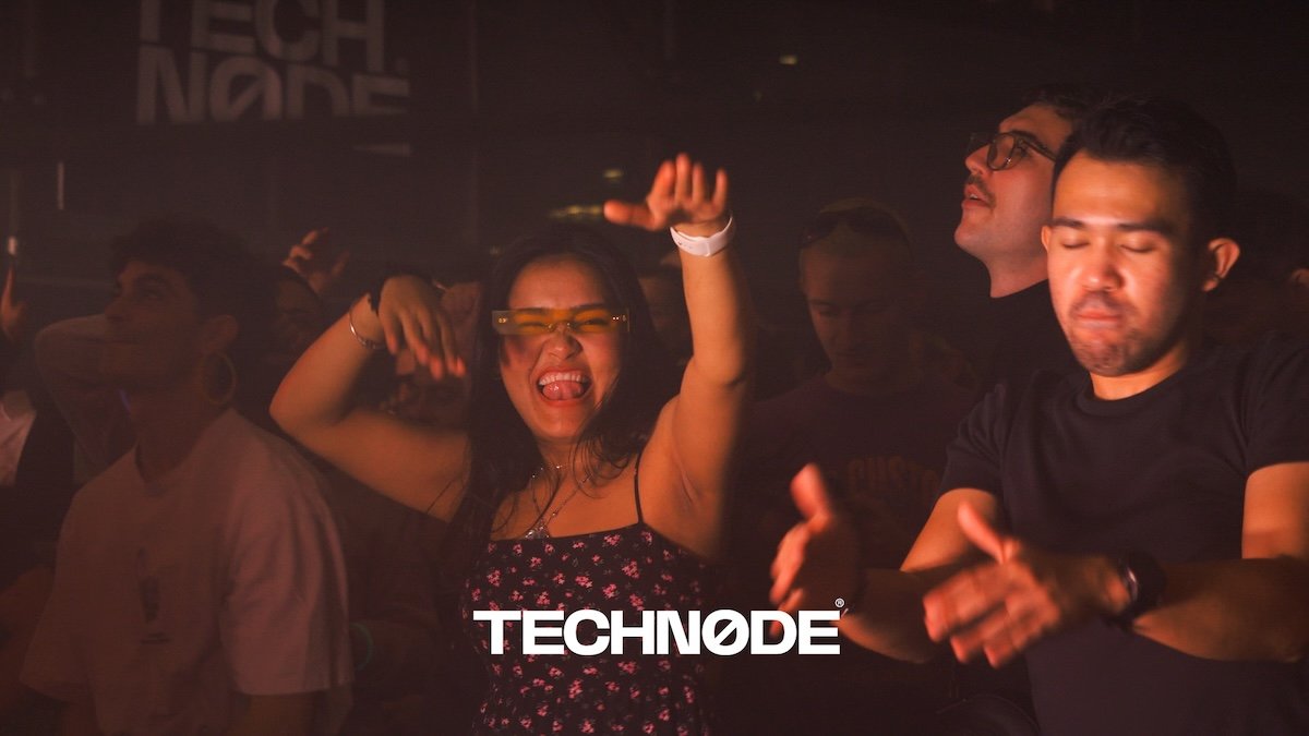 People dancing and enjoying music at a nightclub or concert, with a woman in the center smiling and wearing glasses, and the word "TECHNO" displayed at the bottom.