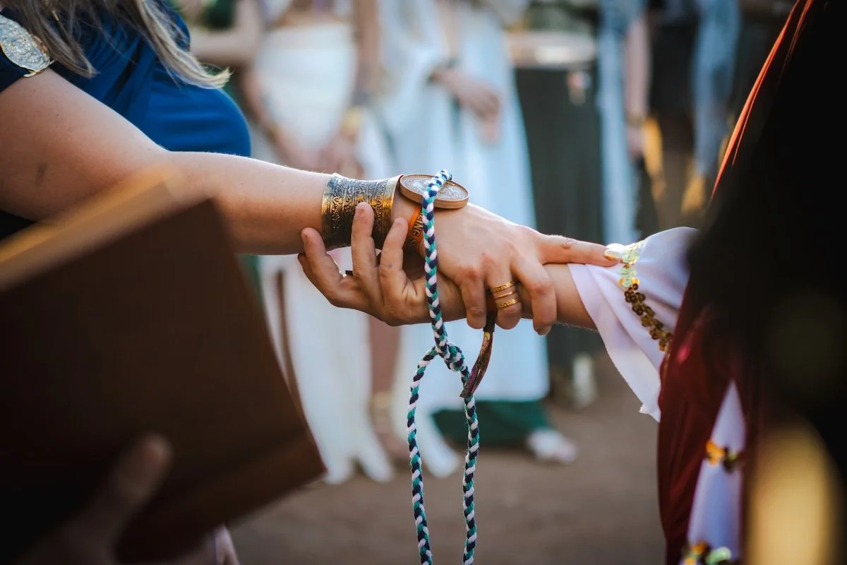 A close-up of two hands clasped during what appears to be a wedding, with one wearing traditional attire and jewelry, and the other hand holding a book. The background shows people dressed in formal or traditional clothing, indicating a ceremonial or