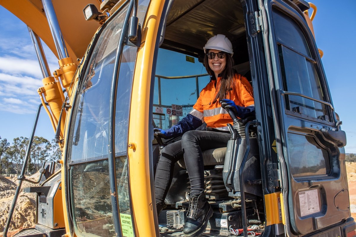 A smiling woman wearing safety gear, including a white hard hat and orange safety vest, sits in the operator's seat of a yellow excavator on a construction site.