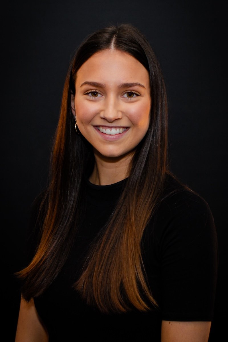 A young woman with long straight brown hair smiling at the camera, wearing a black top, against a black background.