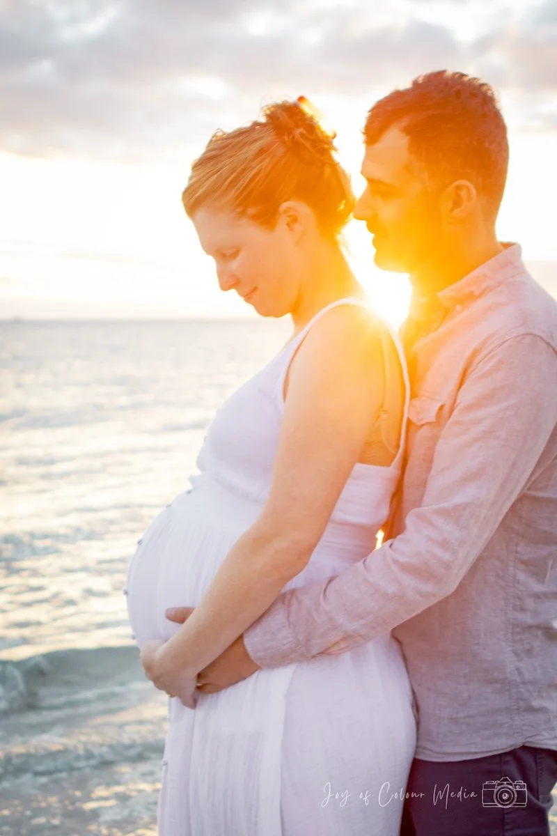 A pregnant woman and a man standing on the beach during sunset, with the man holding the woman from behind. The woman is looking down at her belly, and the man is looking at her. The sunlight creates a warm glow around them.