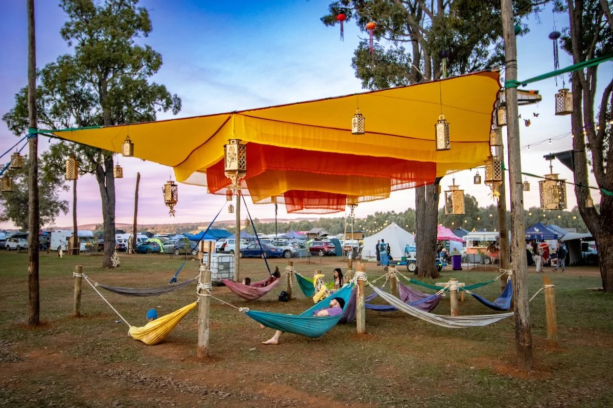 People relaxing in hammocks under a canopy at an outdoor festival with tents, cars, and trees in the background during sunset.