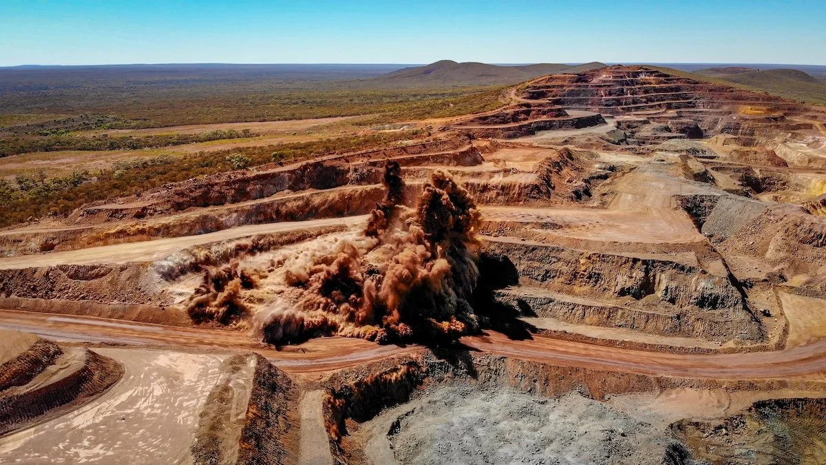 An open-pit mine with a large dust cloud rising from an excavation site, surrounded by layers of earth and sparse vegetation in the background.