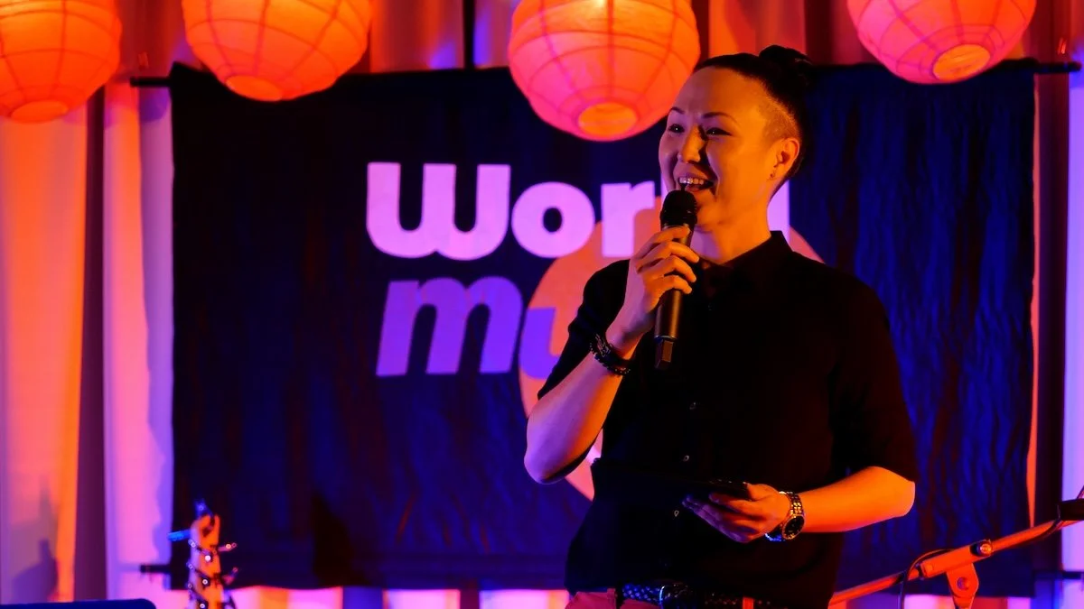 A person with short hair holding a microphone and smiling on stage, with orange paper lanterns hanging overhead and a black banner with logo and text in the background.