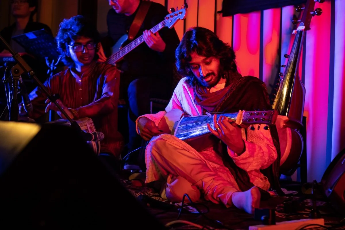 Musicians performing on stage with traditional Indian musical instruments, including a seated man playing a sitar and a boy playing a tabla, under colorful stage lighting.