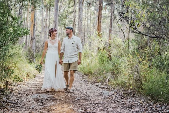A couple walking hand in hand on a dirt trail in a forest, with the woman wearing a white dress and the man dressed casually in a shirt and shorts.