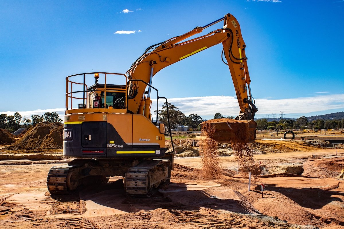 An orange excavator at a construction site, scooping dirt with its bucket, with a partly cloudy sky and distant trees and hills in the background.