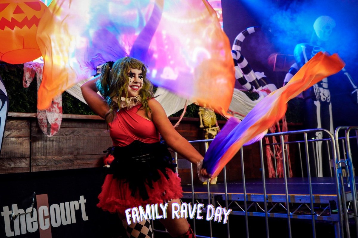 A woman with curly hair and clown makeup dances with colorful fabric at an event called 'Family Rave Day,' decorated with Halloween-themed balloons and skulls.