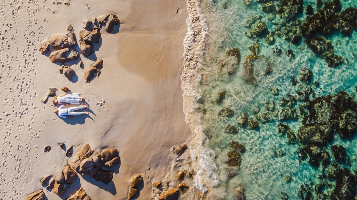 Aerial view of a beach with rocks, sand, and clear blue water, showing a couple lying on the sand.