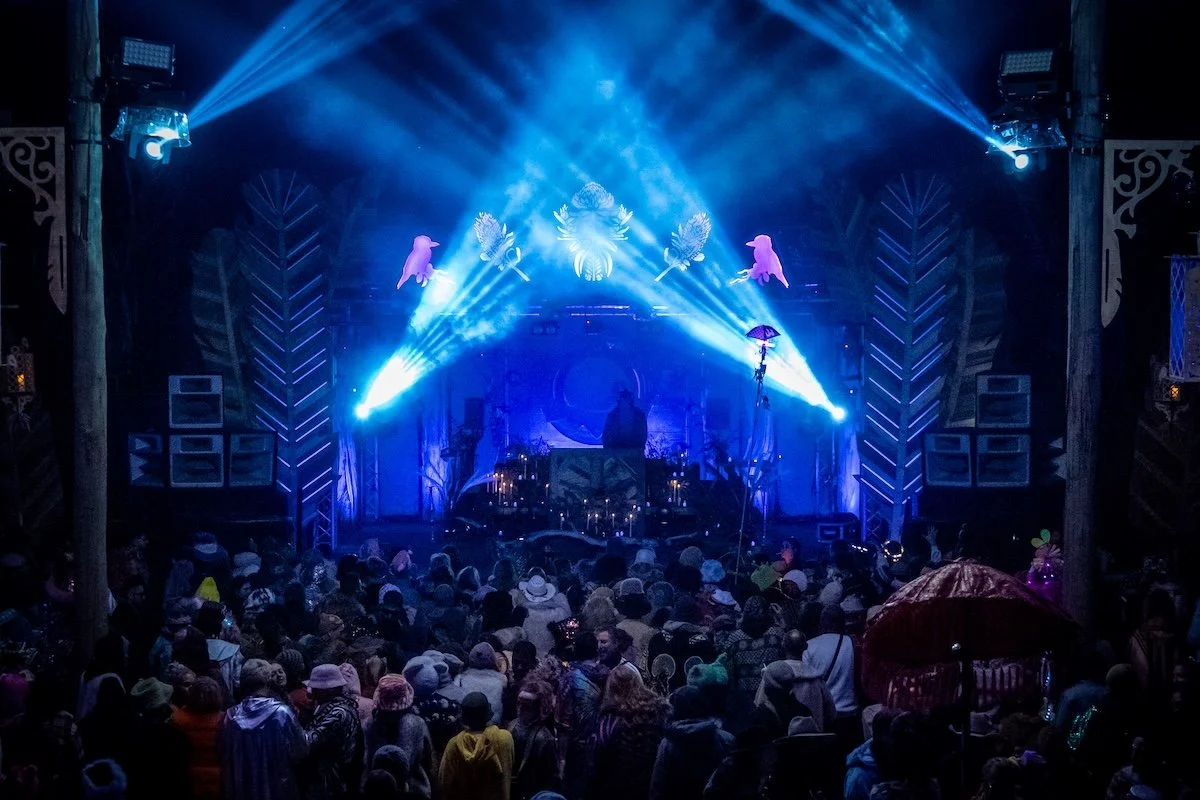 A live music concert on a stage with blue lighting, decorative wall art, and a crowd of people watching, some holding umbrellas.
