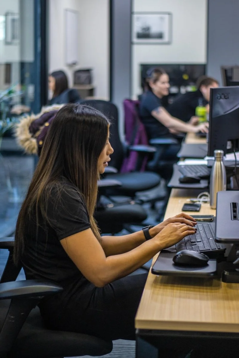 Woman working on a computer in an office with several colleagues in the background.