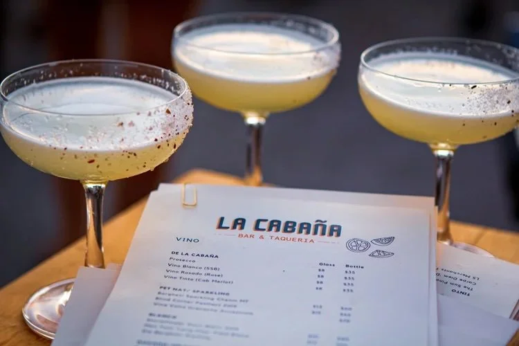 Three cocktails in coupe glasses on a table with a La Cabaña bar and taqueria menu.