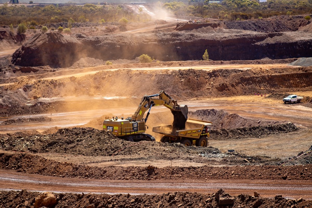 Excavator loading dirt into a dump truck in a large open pit mining site with multiple tiers of earth and machinery.