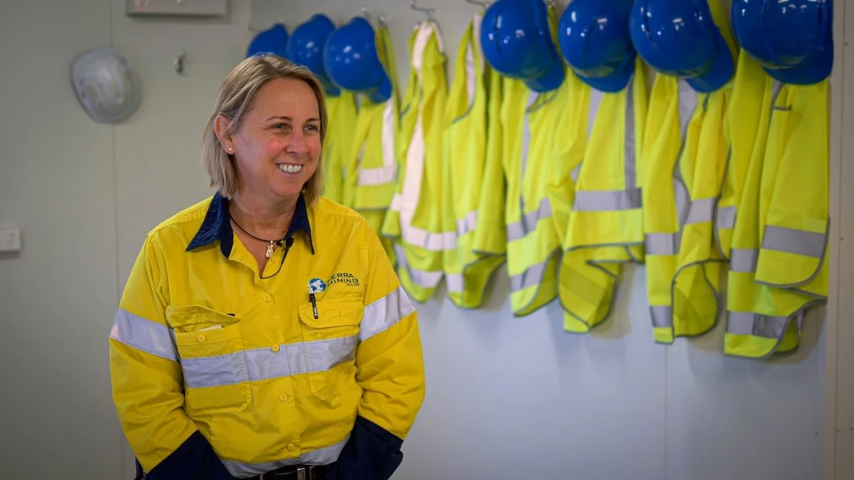 A woman in a yellow safety jacket standing in front of a row of yellow safety vests and blue helmets hanging on a wall.