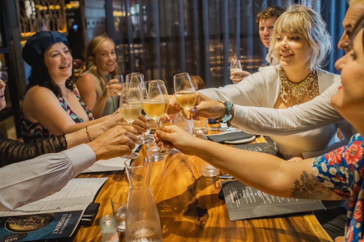 People around a table raising glasses for a toast with white wine, smiling in a celebratory setting.