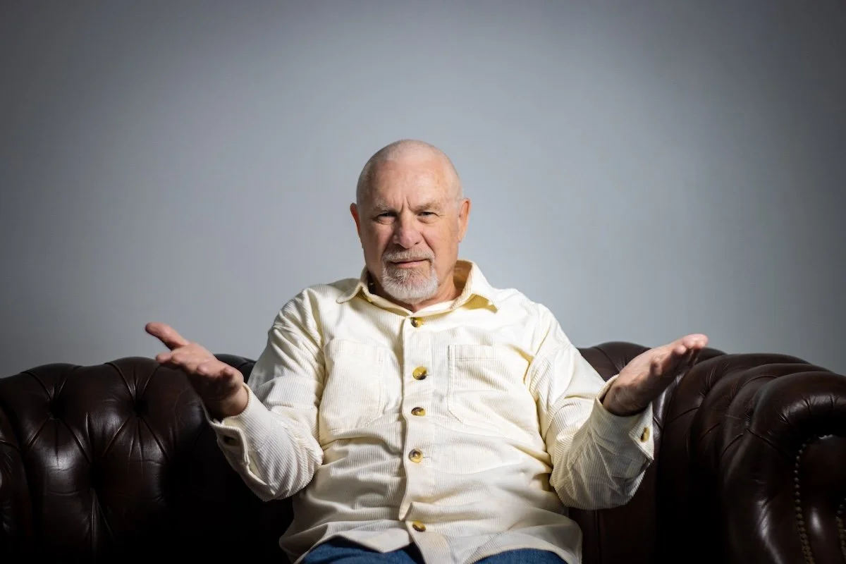 Older man with a confused or questioning expression, gesturing with palms up, sitting on a brown leather sofa against a plain gray background.
