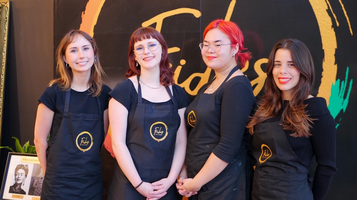 Four women standing side by side, wearing black aprons with a yellow circular logo, smiling indoors against a black background with colorful text and graphics.