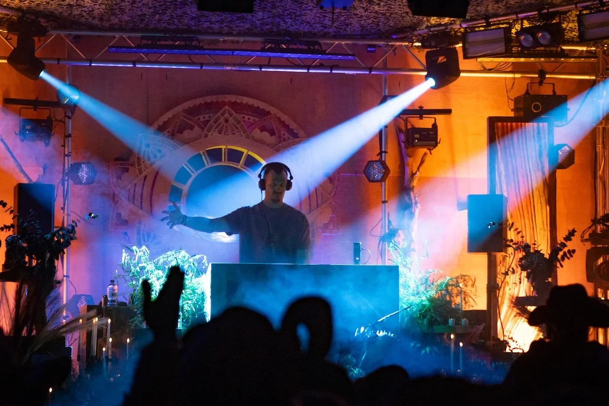 A DJ is performing on stage with colorful lights and smoke effects, with a crowd watching in front.