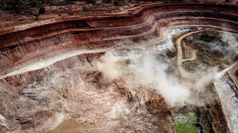 A large open-pit mining site with multiple terraced levels, smoke rising from the excavated area, and a winding dirt road leading into the mine.