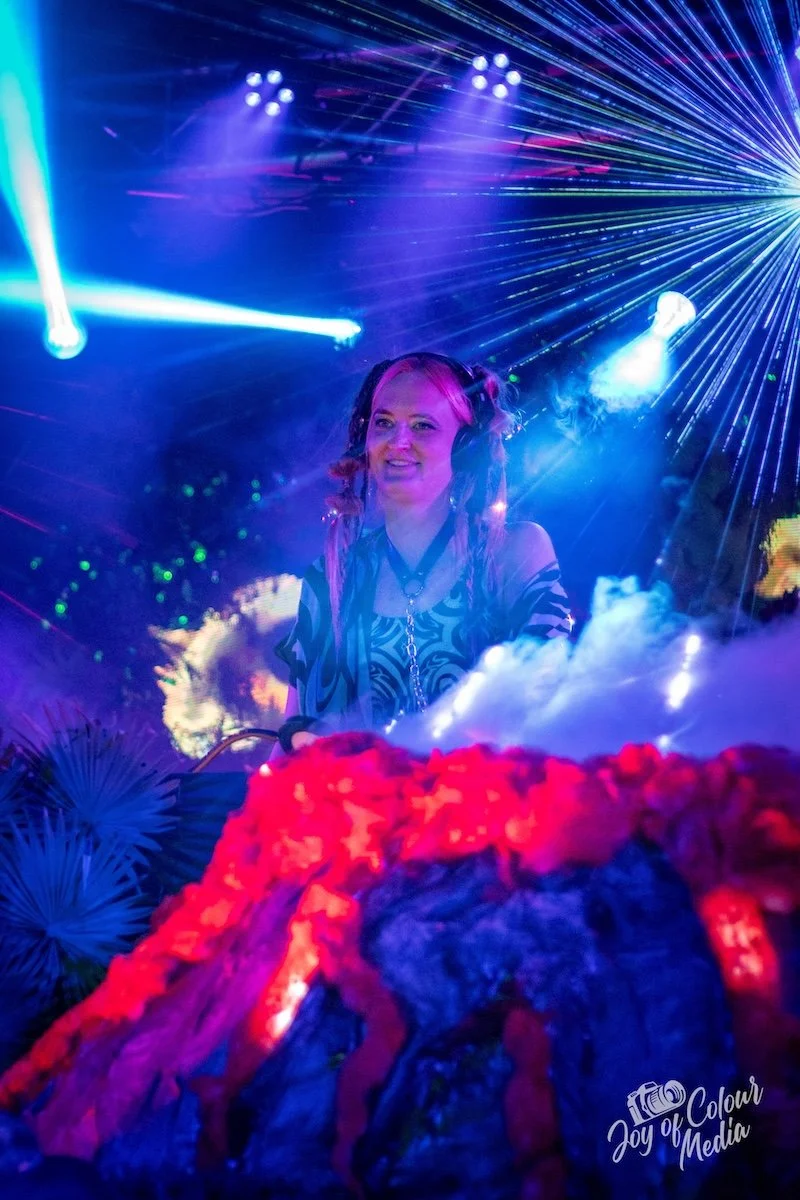 A female DJ with pink and purple hair wearing headphones and a patterned top, smiling as she performs at a nightclub with colorful laser lights and fog.
