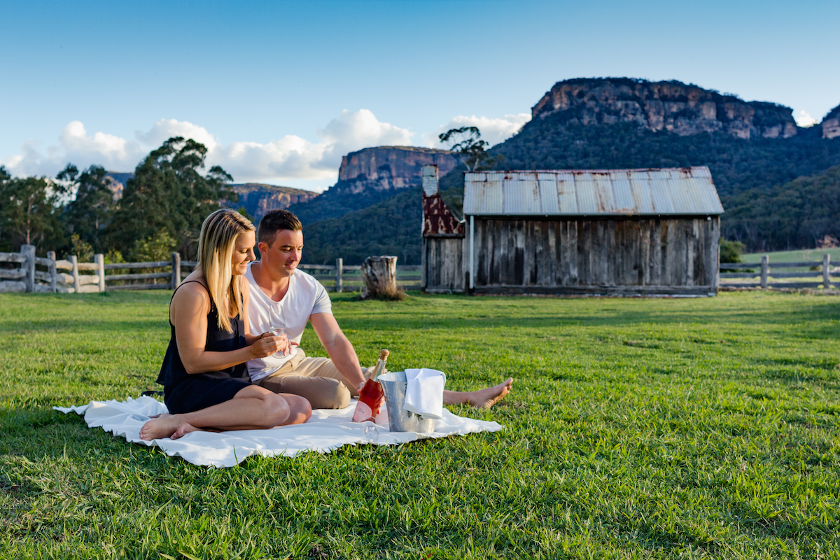 A couple having a picnic on a white blanket in a rural field, with an old wooden barn and mountains in the background.