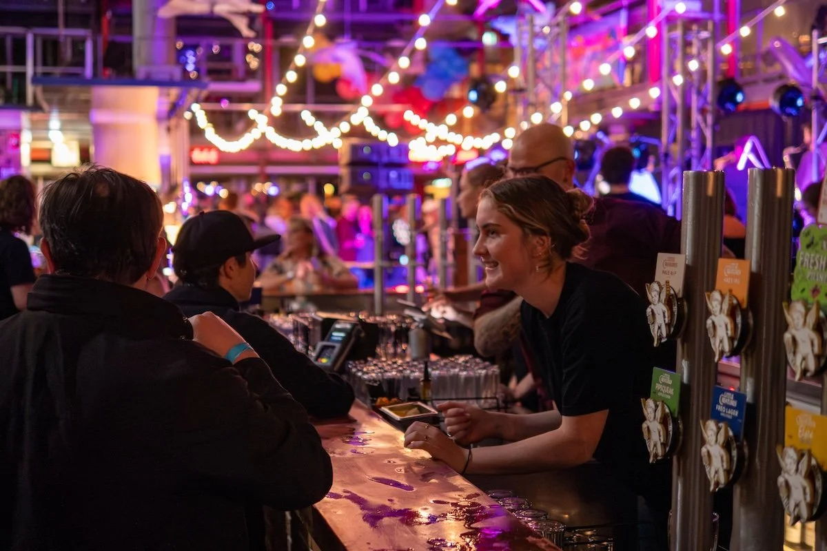 People at a lively nightlife bar with colorful lights, a bartender smiling, and a crowded background.