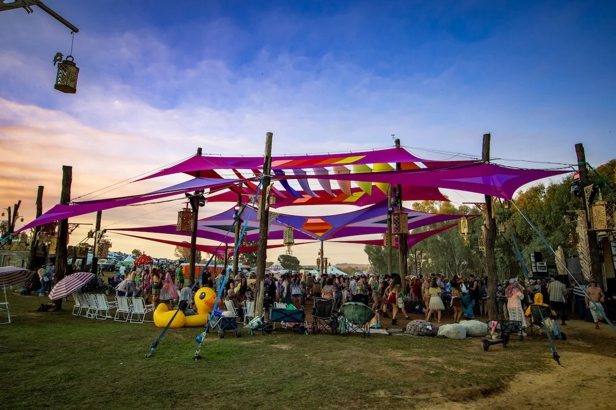 An outdoor festival scene at sunset with a colorful fabric canopy, groups of people, chairs, and decorations hanging from wooden poles.