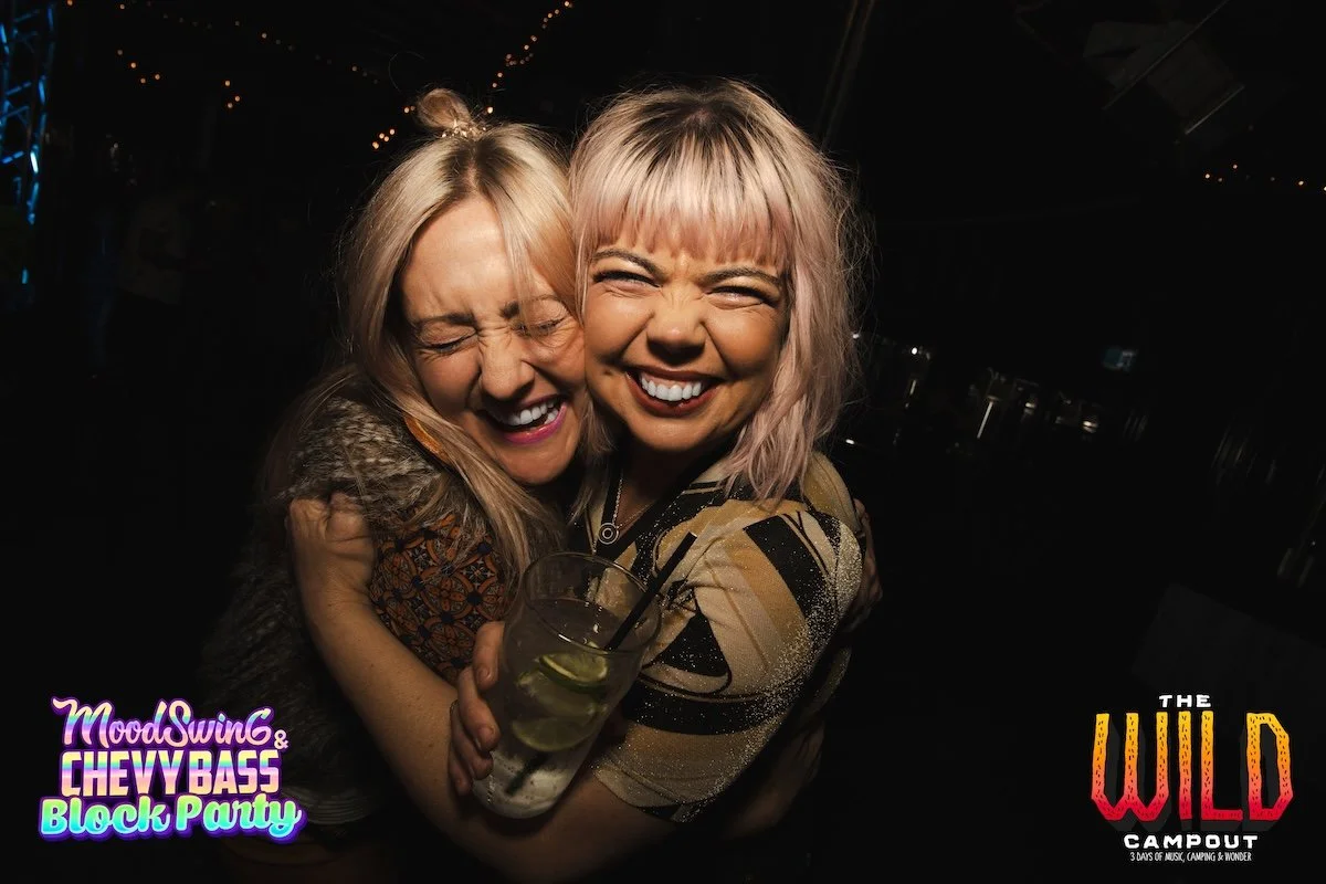 Two women hugging and smiling at a lively party. One woman is holding a drink with lime and straw. The background is dark with string lights, indicating a night event.