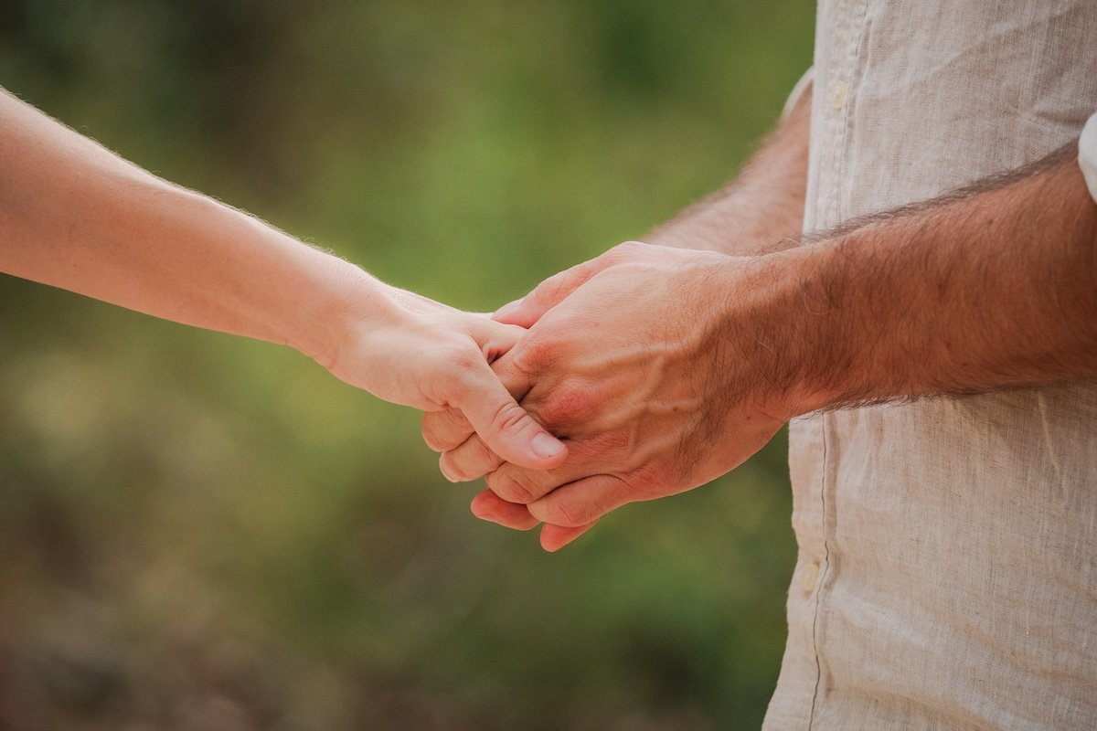 Close-up of two people holding hands, outdoors with a blurred green background.