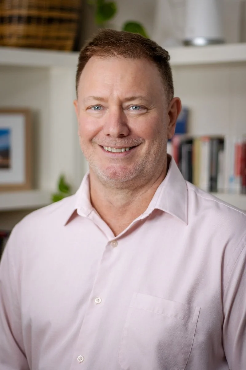 A middle-aged man with short brown hair and blue eyes, smiling, wearing a light pink dress shirt, standing in a room with bookshelves and framed pictures.