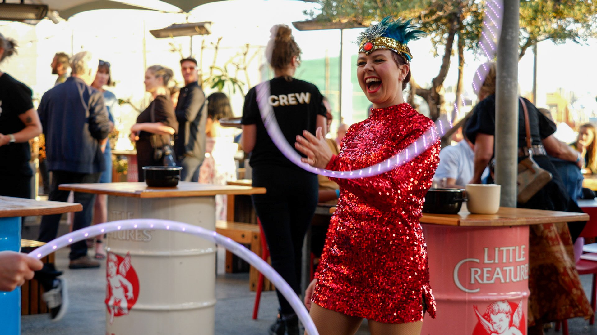 A woman in a red sequined dress and colorful feathered headpiece dancing and smiling at an outdoor event, with people socializing and music equipment in the background.
