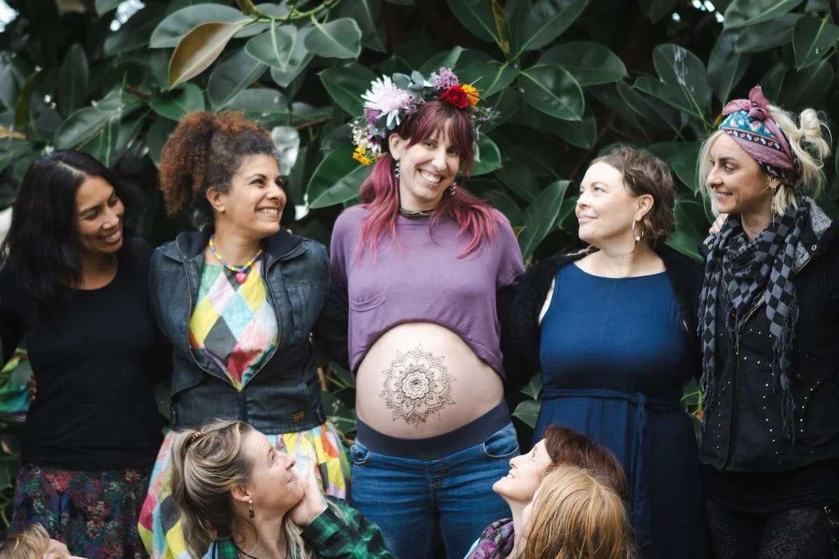 Group of women smiling together outdoors, celebrating a woman with a floral crown and a mandala tattoo on her belly, surrounded by greenery.