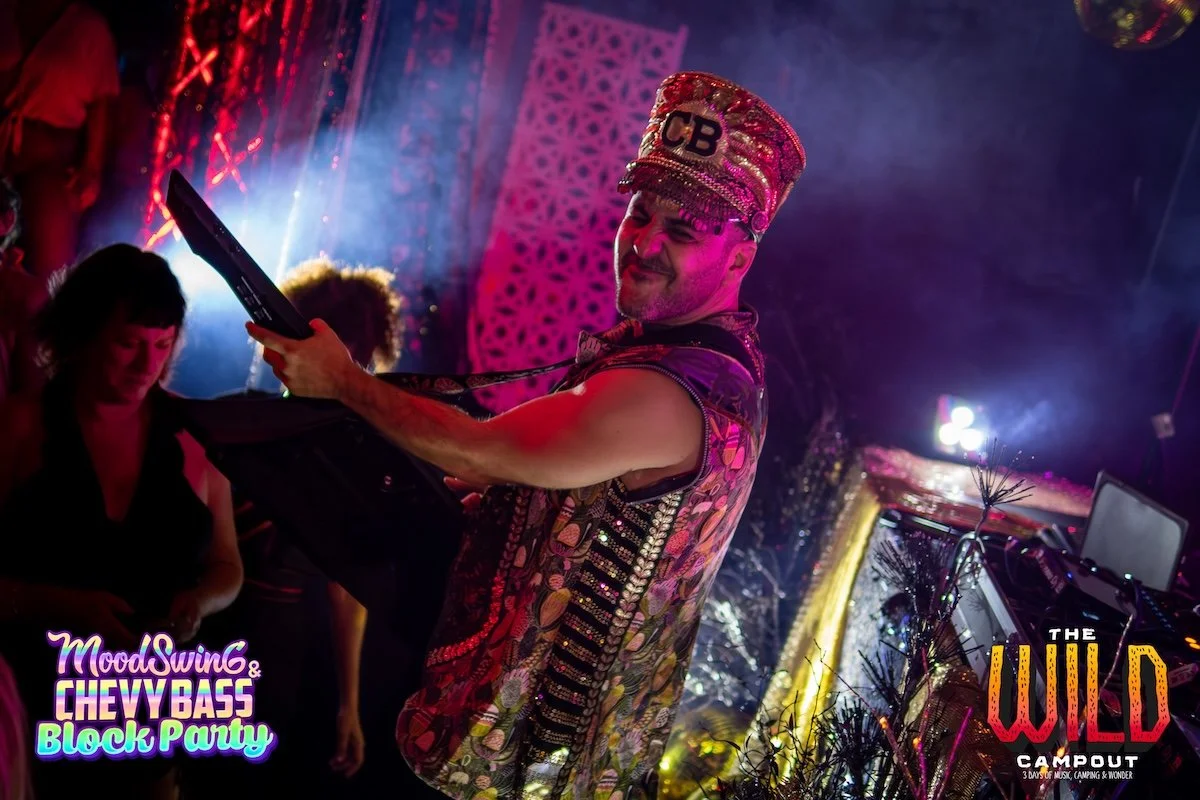 A DJ wearing a colorful vest and a decorated hat with "CB" on it performs at a party with colorful lighting and other people dancing in the background.