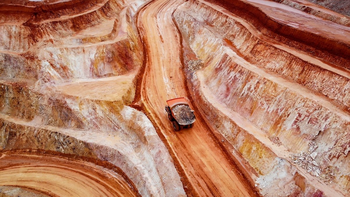 An orange dump truck hauling a load of dirt on a winding dirt road inside a colorful open-pit mine with terraced rock formations.