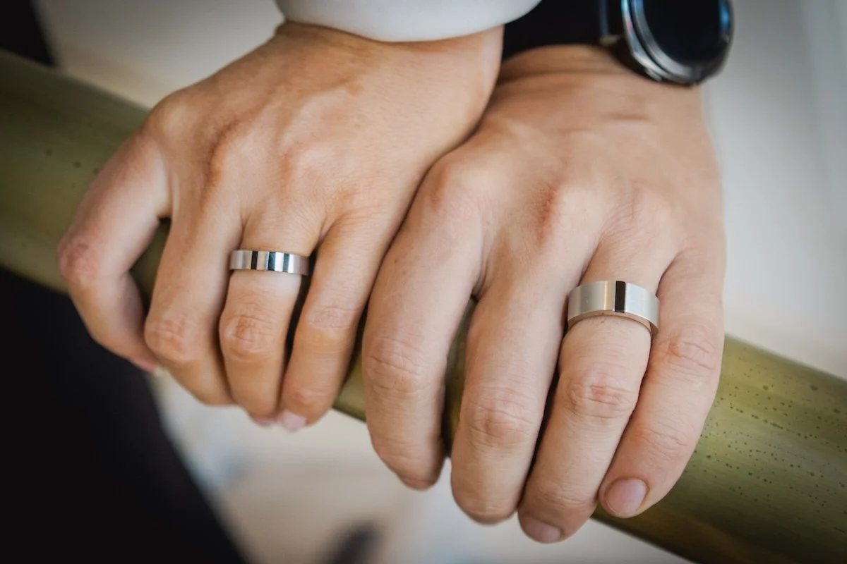 Two hands clasped over a wooden surface, each wearing a matching silver ring.