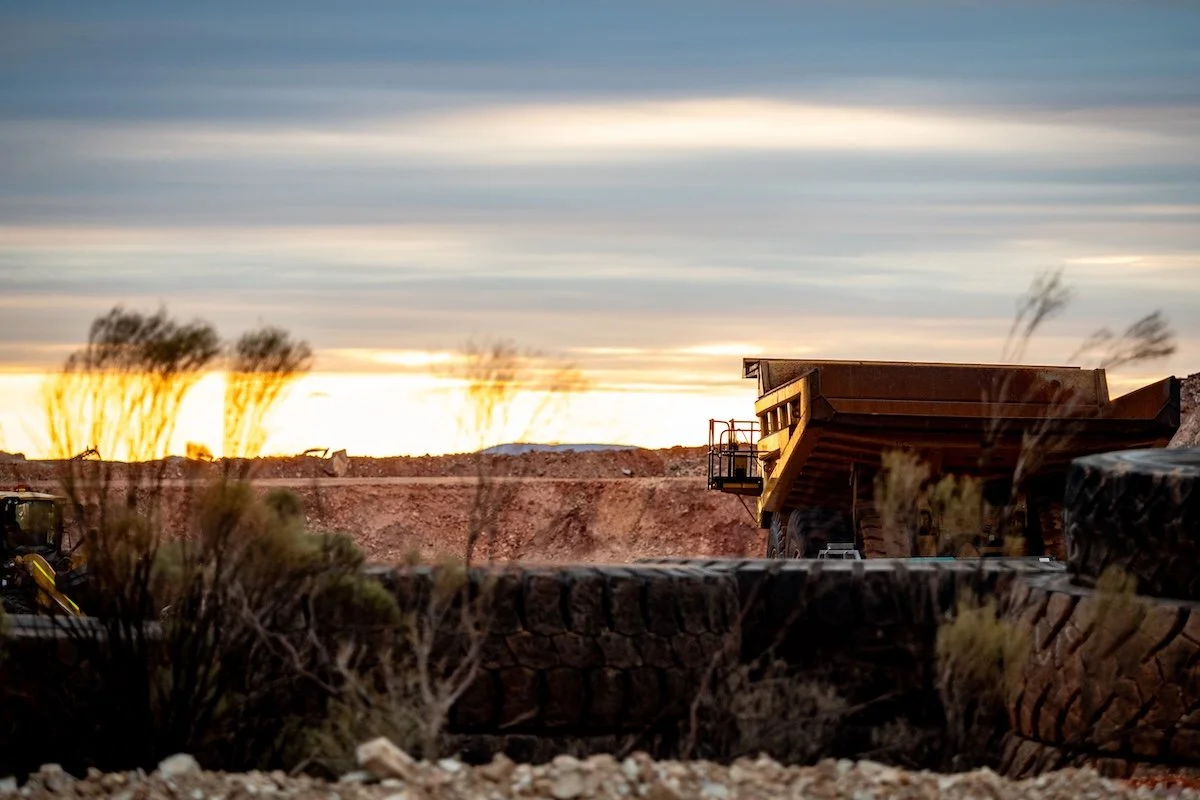 Construction site during sunset with a large dump truck, a stone wall, and desert-like terrain with sparse vegetation.