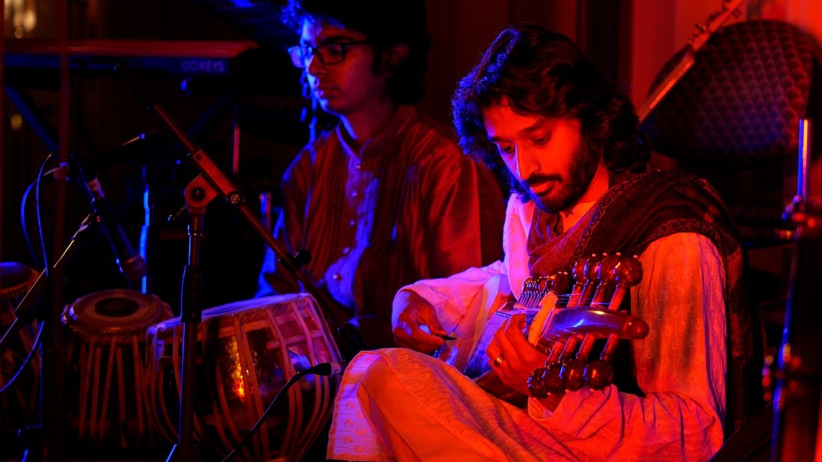 Two musicians sitting in a dimly lit studio, one playing a sitar and the other sitting behind a tabla, illuminated by red and blue lighting.