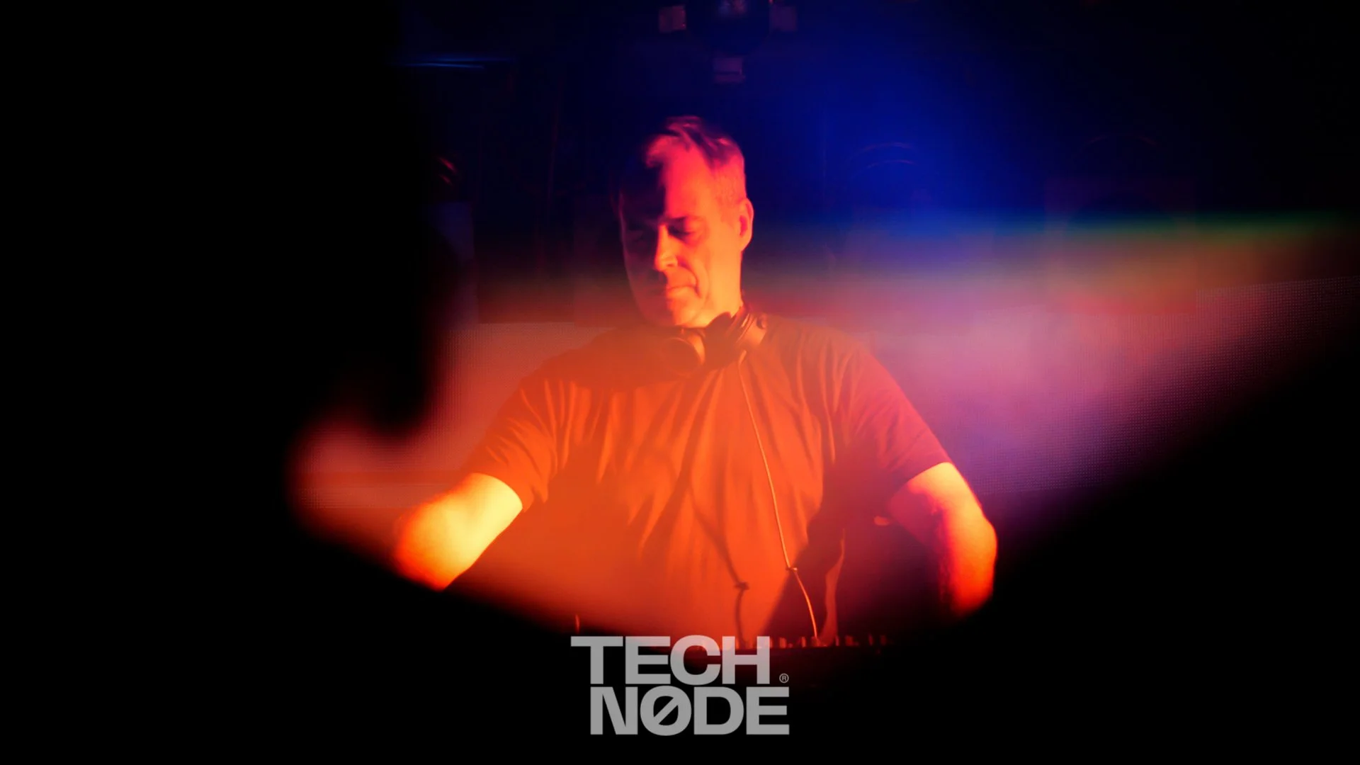 A DJ performs at a Techno event with colorful stage lighting, including red, purple, and blue lights, and the 'TECH NODE' logo at the bottom.