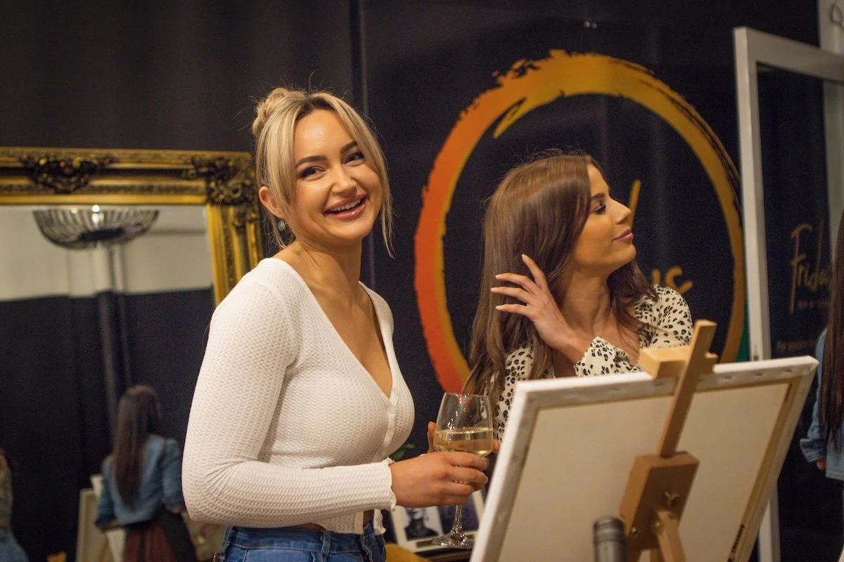 Two women at an art gallery, one smiling and holding a glass of white wine, the other looking at a painting on an easel.