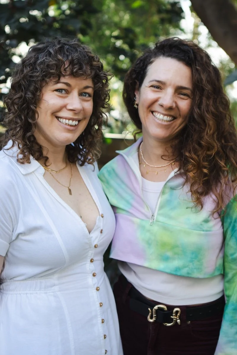 Two smiling women outdoors, one with curly brown hair and the other with wavy brown hair, standing close together with a blurred green background.