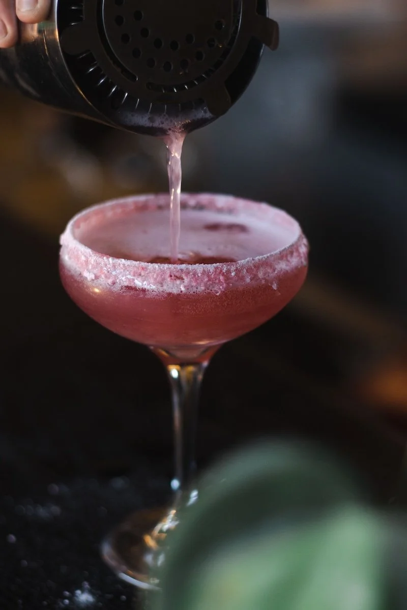 A pink cocktail in a fancy glass with a frosted rim, being poured from a cocktail shaker.