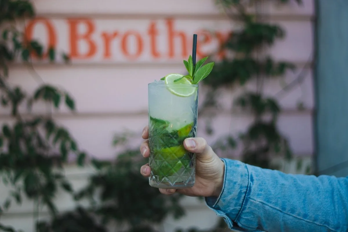 A person holding a refreshing mojito cocktail garnished with lime slices and mint leaves, with a straw, in front of a blurred background with a sign that says 'O Brother.'