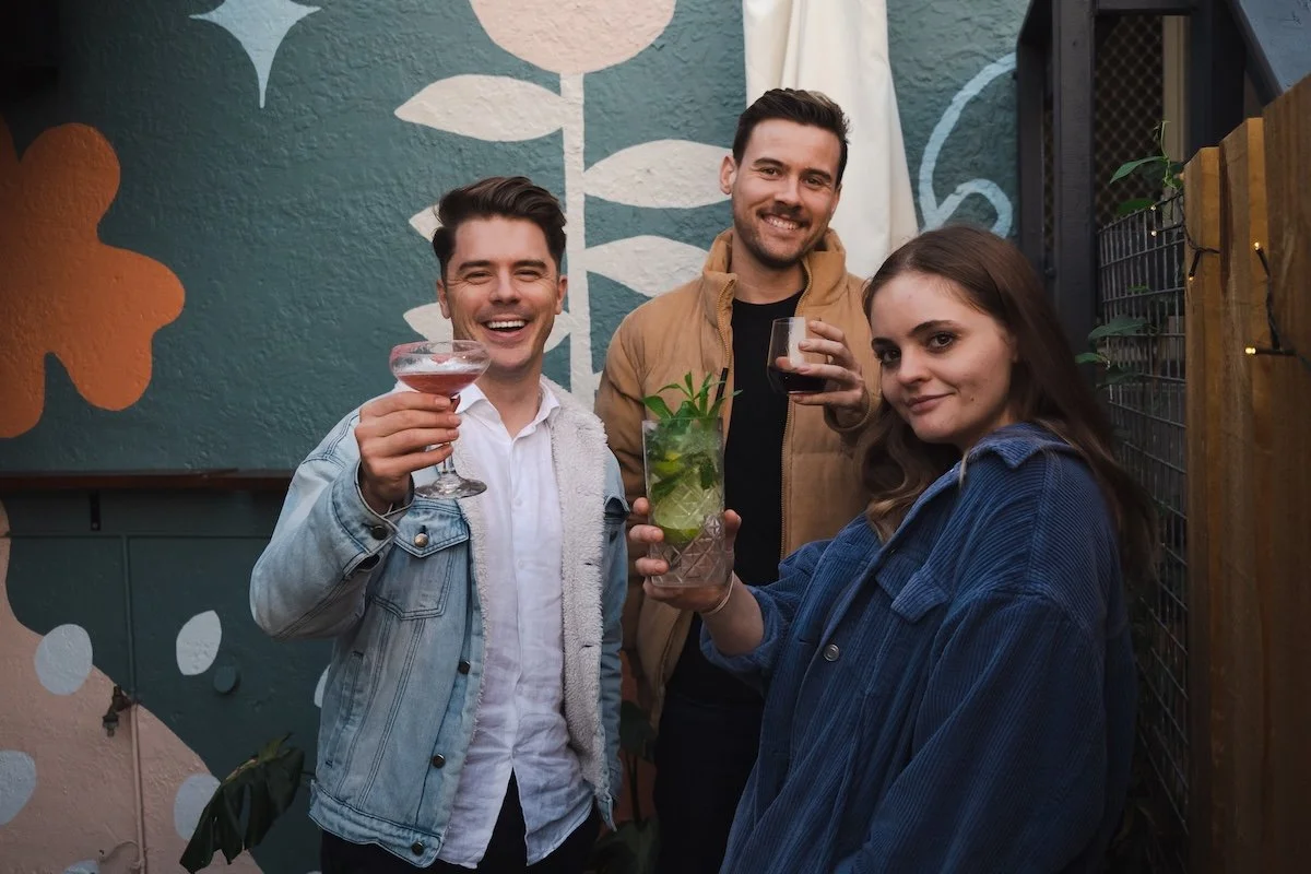 Three friends smiling and holding drinks at a backyard gathering during the evening, standing in front of a colorful painted wall and wooden fence.