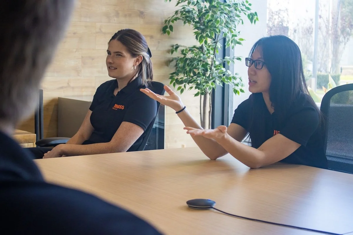 Two women in black shirts seated at a conference table, one gesturing while speaking, the other listening, with a man partially visible in the foreground, in a bright room with large windows and a potted plant in the background.